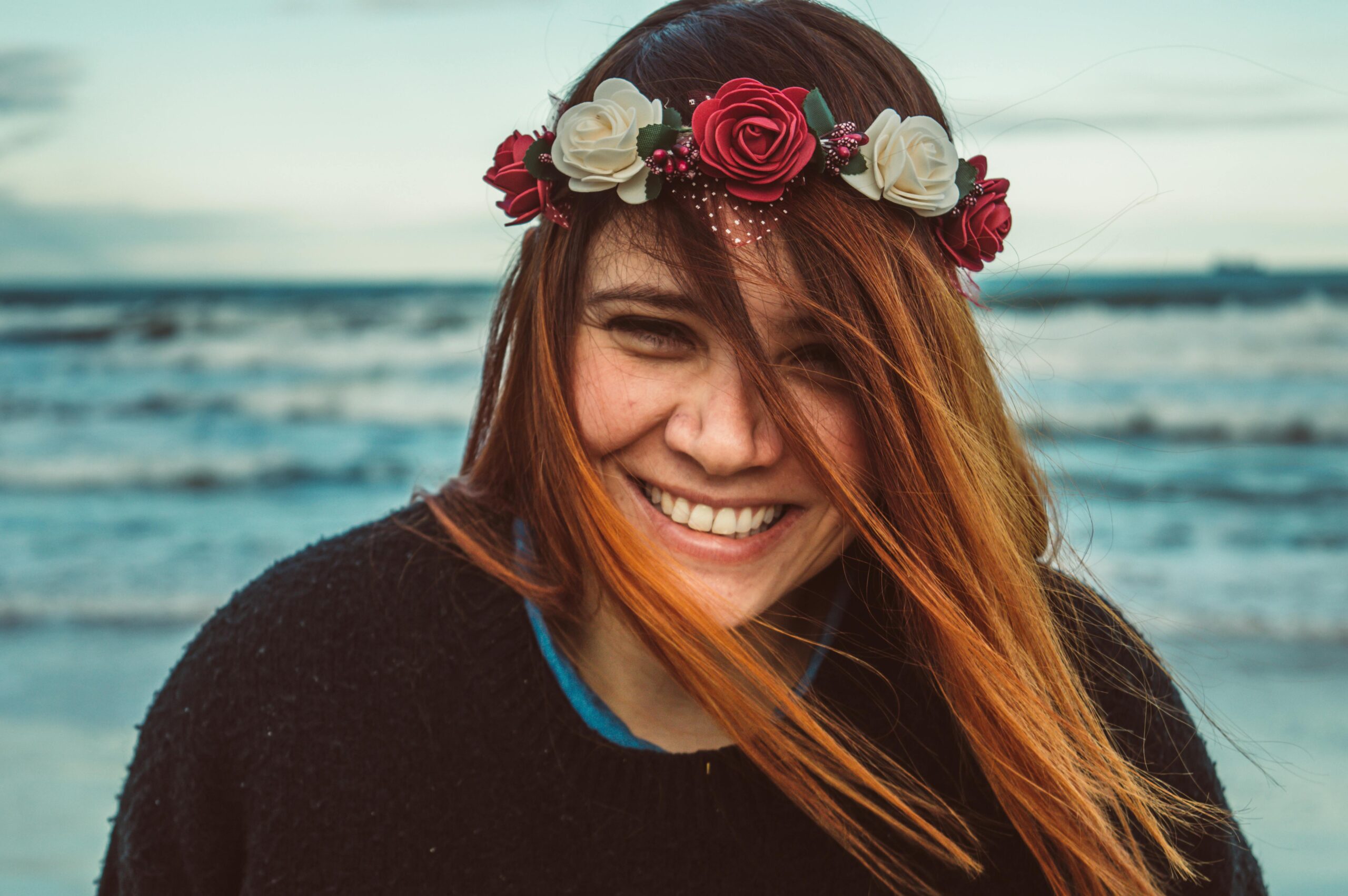 A woman smiling with a flower crown, enjoying a breezy day at the beach.